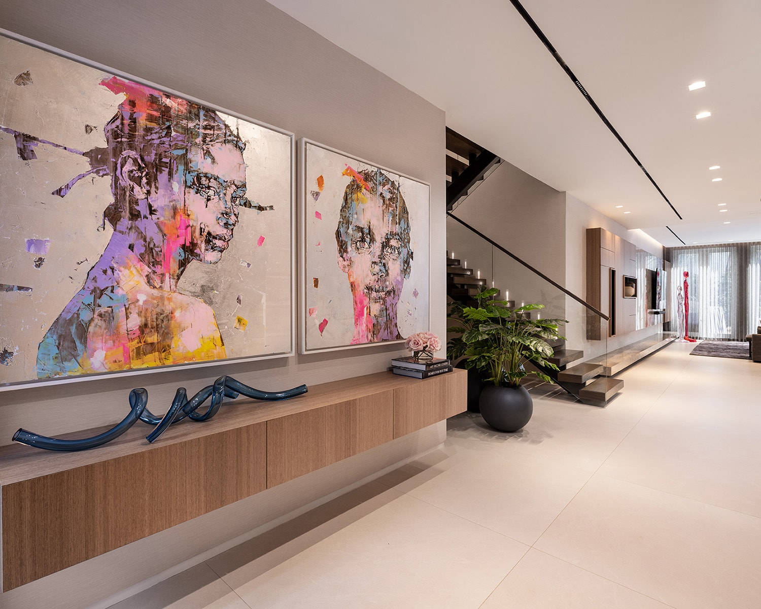 A modern, luxury home corridor showcasing bespoke art and styling. Features large, vibrant contemporary portraits above a floating wood console, a sleek open staircase, integrated linear ceiling lighting, and a long, minimalist hallway leading to a bright living space.